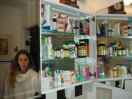 A white medicine cabinet with multiple shelves. On the left side of the cabinet there is a woman with long brown hair wearing a white sweater. She appears to be deep in thought with her eyes closed and her head resting on her hand. The shelves are filled with various medical supplies such as bottles containers and containers of different sizes and colors. There is also a clock on the wall above the cabinet. The cabinet is open revealing the contents inside.