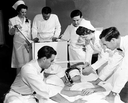 A black and white photograph of a group of medical professionals gathered around a patient lying on a hospital bed. The patient appears to be unconscious and is covered with a white sheet. There are six people in the image all wearing white uniforms and caps and one person is holding a clipboard. They are gathered around the bed looking at the patient with concern. One person is kneeling on the bed and another is standing next to the bed. Another person is standing behind the bed with their hands on the patient's head. There is a desk with a computer monitor and other medical equipment in the background. The overall mood of the image is somber and somber.