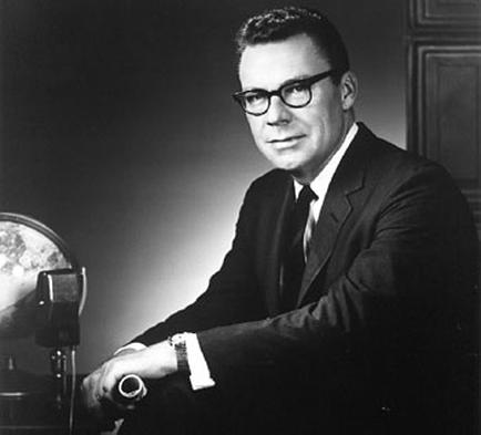 A black and white portrait of a man sitting at a desk with a globe in front of him. The man is wearing a suit and tie and has a serious expression on his face. He is wearing glasses and appears to be in his late 40s or early 50s. The desk is made of wood and there is a lamp on the right side of the image. The background is dark and the lighting is dim creating a moody atmosphere.