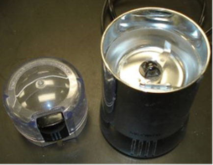 Two objects on a black surface. On the left side there is a small plastic container with a lid. The lid is transparent and appears to be made of plastic. The container on the right side is black and has a cylindrical shape with a handle on top. It has a small hole in the center which is likely for the lid to be inserted into the container. The background is blurred but it seems to be a kitchen countertype.