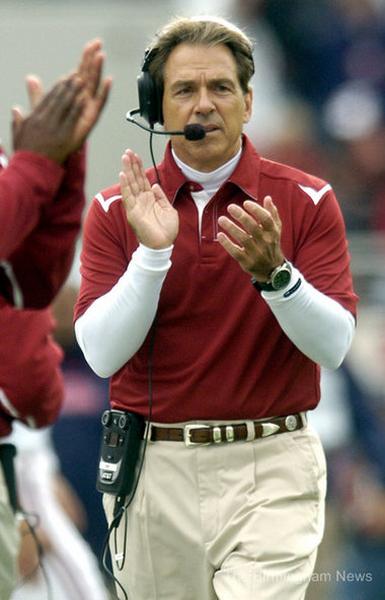A man wearing a maroon polo shirt and beige pants standing on a football field. He is wearing a headset with a microphone attached to it and appears to be a coach or a member of the football team. He has a serious expression on his face and his hands are raised in the air as if he is clapping or cheering. In the background there are other players and spectators in the stands. The image appears to have been taken during a football game.