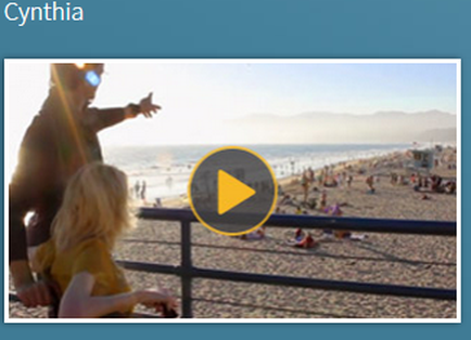 A man and a woman on a beach. The man is standing on the left side of the image pointing towards the ocean with his right hand. He is wearing a black t-shirt and sunglasses. The woman is sitting on the ground in front of him looking out at the beach. In the background there are many people on the beach some sitting and some standing. The beach is sandy and there are mountains in the distance. The sky is blue and the sun is shining brightly. There is a yellow play button in the bottom right corner of the screen.