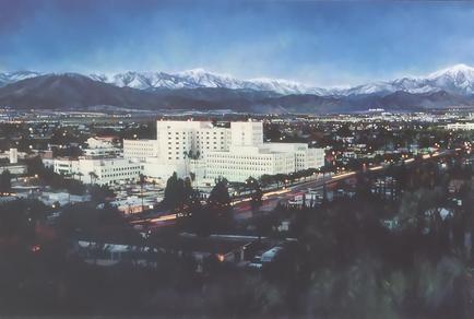 An aerial view of a city with a large white building in the center. The building appears to be a hotel or a conference center with multiple floors and multiple windows. It is surrounded by trees and other buildings and there are several roads and highways visible in the foreground. In the background there are snow-capped mountains and a clear blue sky. The image is taken from a high vantage point giving a bird's eye view of the city below.