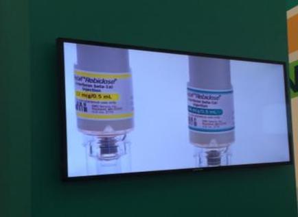 A flat-screen TV mounted on a green wall. On the TV screen there are two syringes one with a white cap and the other with a blue cap. The syringe on the left has a label that reads "sterile-Rubbicide" and the label on the right has the same label as the one on the bottom. The label also has the words "1 ml/0.5 ml" written on it. The TV screen appears to be displaying a product or advertisement.