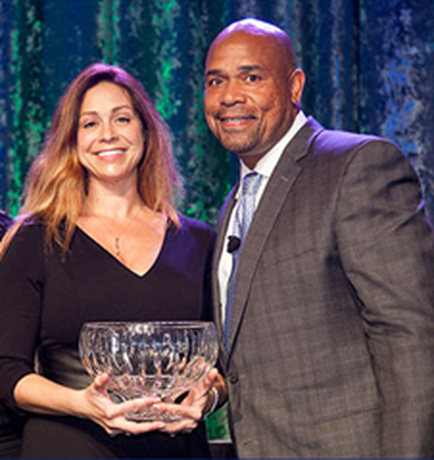 A man and a woman standing on a stage with a blue curtain in the background. The woman is on the left side of the image and is holding a large crystal bowl in her hands. She is wearing a black dress and has long blonde hair. The man on the right side is also wearing a suit and tie. They are both smiling and looking at the camera. It appears that they are posing for a photo.
