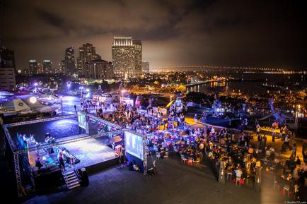 An aerial view of a large outdoor event taking place at night. The event is taking place on a rooftop with a city skyline in the background. The sky is dark and cloudy and the city is lit up with colorful lights. In the foreground there is a stage set up with a band performing on it. The stage is surrounded by a crowd of people and there are several airplanes parked nearby. The city skyline is visible in the distance with tall buildings and skyscrapers. The water below is calm and reflects the lights of the city. The overall mood of the image is lively and energetic.