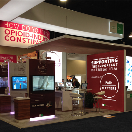 A trade show booth with a red and white color scheme. The booth has a large banner that reads "How do you treat opioid-induced constipation?" and a sign that says "Supporting the important role we each play." There is also a desk with a computer monitor and a chair in front of the booth. In the background there are other booths and people walking around. The overall atmosphere of the image is professional and inviting.