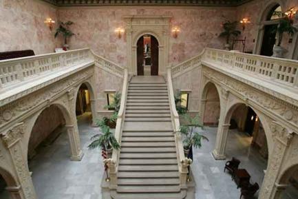 A grand staircase in a luxurious building. The staircase is made of stone and has a high ceiling with intricate carvings and designs. The walls are painted in a light beige color and there are several arches on either side of the staircase. The arches are decorated with potted plants and there is a large archway at the top of the stairs leading to the second floor. The floor is covered with a patterned rug. The overall atmosphere of the space is elegant and opulent.