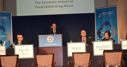 A panel discussion taking place at a conference. There are four people sitting at a table with microphones in front of them. The table is covered with a blue tablecloth and there are nameplates on the table. On the left side of the image there is a man standing behind a podium with a microphone and speaking into it. He is wearing a suit and tie and appears to be giving a speech. Behind him there are two banners with the text "The Economic Impact of Prescription Drug Abuse" and a logo of the United States Department of Health and Human Services. The background is a large screen with the same text and logo.