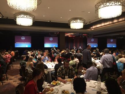 A large conference room with a high ceiling and chandeliers hanging from it. The room is filled with people sitting at long tables facing a stage where a man is giving a presentation. The audience is seated in rows of chairs facing the stage and there are two large screens on either side of the stage displaying the logo of a company or organization. The tables are covered with white tablecloths and have plates of food and drinks on them. The people in the room appear to be engaged in the presentation with some people standing at the front of the room and others seated at the back.
