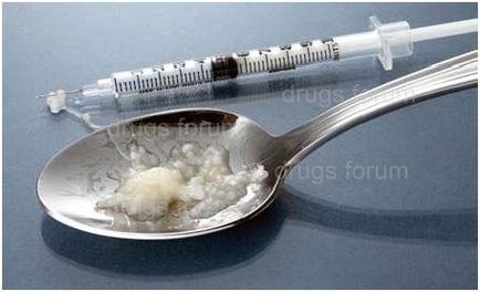 A syringe and a spoon with a small amount of white substance on it. The syringe is on the left side of the image and the spoon is in the center. The substance appears to be a mixture of white and yellow colors possibly sugar or cream. The spoon is silver and has a curved handle. The background is a plain blue color.