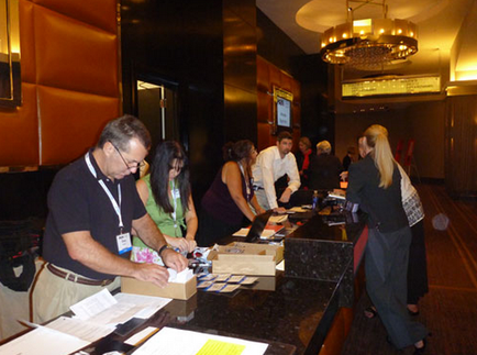 A group of people gathered around a long counter in a lobby or reception area. The counter is covered with papers and other items and there are several people standing around it. A man in a black shirt is signing a document at the counter while a woman in a green shirt is looking at it intently. There are other people in the background some of whom are also signing the document. The lobby has a high ceiling with a chandelier hanging from it and the walls are painted in a warm orange color. There is a large screen on the wall behind the counter and a door on the left side of the image.