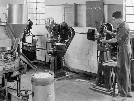 A black and white photograph of a man working in a factory. He is standing in front of a large machine with a large hopper on the left side and a smaller machine on the right side. The man is wearing overalls and appears to be working on the machine. In the background there are other machines and equipment scattered around the room. The floor is tiled and there is a large window on the far wall. The image appears to have been taken in the early 20th century.