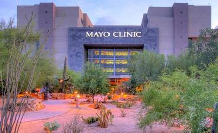 The exterior of a Mayo Clinic building at dusk. The building is a modern two-story structure with a gray exterior and large windows. The word "Mayo Clinic" is written in large white letters on the top of the building. The sky is a deep blue and the building is lit up with warm orange and yellow lights. In front of the clinic there is a pathway lined with cacti and succulents and a few people can be seen walking around. The overall atmosphere of the image is peaceful and serene.