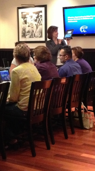 A group of people sitting in a dining room with wooden chairs. There are six people in the image three women and three men all of whom appear to be engaged in a conversation. The woman in the center of the image is standing and holding a tablet in her hand while the other three women are sitting at the table in front of her. On the wall behind them there is a framed black and white photograph of a football game. The room has a large flat-screen TV mounted on the wall and a wooden floor.