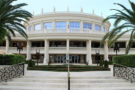 A large luxurious building with a round white exterior and a curved roof. The building has multiple levels with large windows and balconies and is surrounded by palm trees and other tropical plants. In front of the building there is a set of stairs leading up to the entrance. The sky is blue and there are a few clouds in the background. The overall atmosphere of the image is elegant and inviting.
