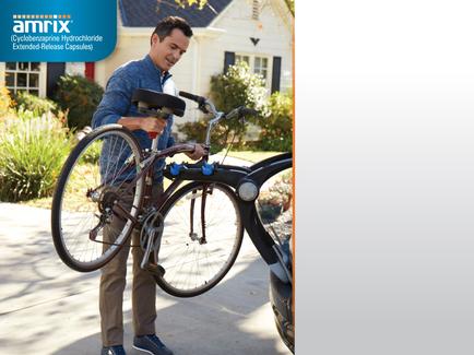 A man standing next to a black Amrix bicycle. He is holding the handlebars of the bicycle with both hands and appears to be fixing it. The man is wearing a blue shirt khaki pants and blue sneakers. He has a smile on his face and is looking down at the bike. The background shows a house with a white exterior and a garden with plants and trees. The Amrix logo is visible in the top left corner of the image.