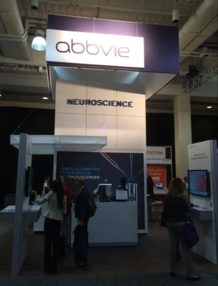 A trade show booth with a purple and white sign that reads "abbvie Neuroscience". The booth is located in a large hall with a high ceiling and white walls. There are several people standing in front of the booth some of them are looking at the sign. On the right side of the image there is a desk with a computer monitor and other equipment. In the background there are other booths and information boards.