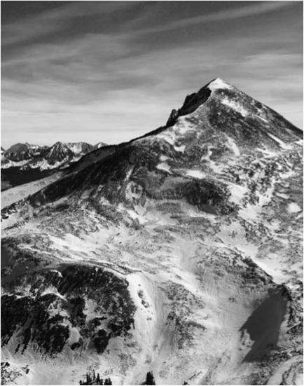 A black and white photograph of a mountain range. The mountain appears to be covered in snow and ice with patches of snow on the peaks and valleys. The sky is cloudy and the mountains in the background are also covered in a layer of snow. The photograph is taken from a high vantage point looking down on the mountain and the surrounding landscape. The overall mood of the image is bleak and desolate.