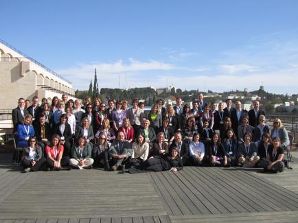 A large group of people posing for a group photo on a wooden deck. There are around 20 people in the photo all dressed in formal attire. They are arranged in a semi-circle with some standing and some kneeling. The deck is located on a hilltop with a clear blue sky and trees in the background. On the left side of the image there is a building with arches and pillars and on the right side there are buildings and a tower visible in the distance. The group appears to be of different ages and ethnicity's and they are all smiling and looking at the camera.