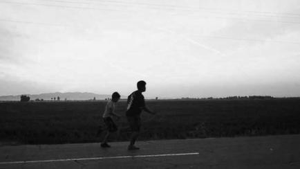 A black and white photograph of two people walking on a road. The road appears to be empty with no cars or other vehicles visible in the background. The sky is overcast and the horizon is visible on the right side of the image. In the distance there are a few buildings and trees scattered across the landscape. The two people in the foreground are walking side by side with one person in the front and the other in the back. They are both wearing casual clothes and appear to be in a hurry. The image is taken from a low angle looking towards the horizon.