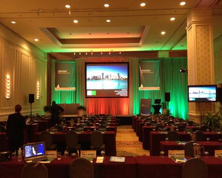 A large conference room with rows of tables and chairs set up for a meeting. The room has a high ceiling with recessed lighting and a chandelier hanging from it. The walls are covered in green curtains and there is a large screen in the center of the room displaying a presentation. On the right side of the screen there are two large screens displaying a city skyline. The tables are covered with red tablecloths and there are nameplates water bottles and laptops on the tables. There are also a few people in the room suggesting that the meeting is taking place in a formal setting.
