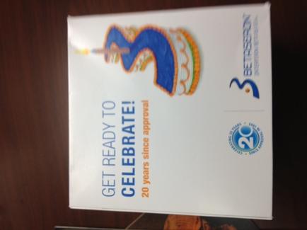 A photograph of a white box with a blue and orange logo on the top right corner. The logo is a stylized letter "S" with a crown on top and the words "Get Ready to Celebrate!" written above it. Below the logo there is text that reads "20 years since approval" in blue. On the bottom right corner of the box there are two blue circles with the number "3" in them. The box appears to be sitting on a wooden surface.