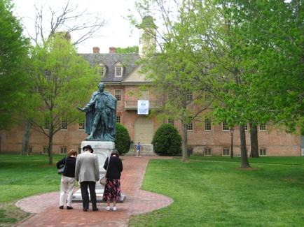 A group of people standing in front of a large brick building with a clock tower. The building appears to be a college or university campus as there are trees and grass surrounding it. In the center of the image there is a statue of a man standing on a pedestal. The statue is of a woman with long hair and a hat and she is holding a book in her hand. The people in the image are looking at the statue and appear to be engaged in conversation. The sky is overcast and the overall atmosphere is peaceful and serene.