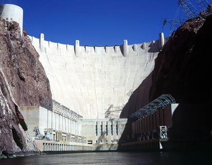 A large dam with a large body of water in front of it. The dam is made of concrete and is located in a canyon-like area with steep cliffs on either side. The walls of the dam are covered in cracks and crevices and there are several large structures on the sides. On the left side of the image there is a tall white tower-like structure which appears to be a hydroelectric dam. The sky is blue and clear and the water is calm and still. The image is taken from a low angle looking up at the dam.