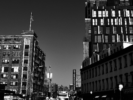A black and white photograph of a city street. The photo is taken from a low angle looking up at the buildings on the left side of the image. The buildings appear to be old and dilapidated with broken windows and crumbling walls. On the right side there is a tall modern building with multiple floors and balconies. The American flag is flying on top of one of the buildings. In the background there are other buildings and a bus on the road. The sky is overcast and the overall mood of the photo is gloomy.