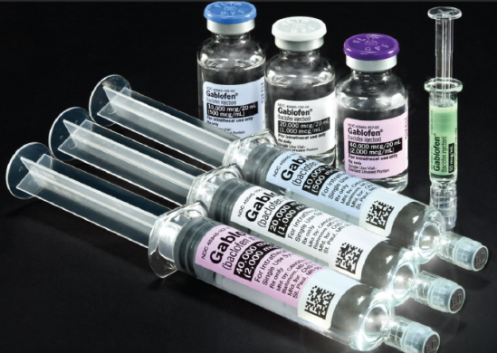A group of four syringes and three vials. They are each labeled with the word "Gablofen" and with information about their prescription strength. Each of the labels is a different color -- blue green white or purple -- based on the dose. The syringes and vials are on a plain black background.