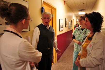 A group of four people in a hospital hallway. On the left side of the image there is a nurse wearing a white coat and a stethoscope around her neck. She is standing in front of a man who is wearing a black vest and glasses. The man appears to be in his 60s or 70s and is gesturing with his hands as if he is explaining something to the two women on the right side of him. The woman on the left is wearing an orange shirt and has curly hair. The other two women are wearing white coats and are also wearing stethoscopes around their necks. They appear to be engaged in a conversation. The hallway has a red door and there are several framed pictures hanging on the walls.
