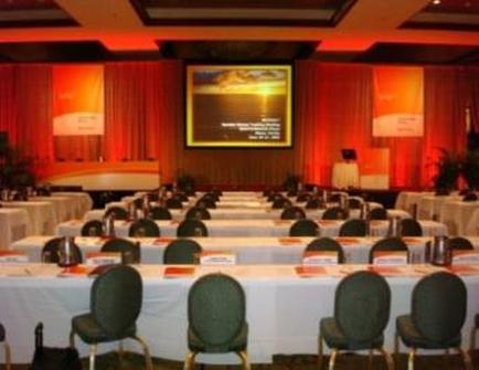 A large conference room with a long table set up for a meeting. The table is covered with white tablecloths and has nameplates and water bottles on it. There are green chairs around the table. The room has red curtains on the walls and a large screen at the front of the room displaying an image of a sunset. On either side of the screen there are two podiums with microphones. The ceiling is high and has recessed lighting.