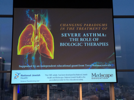 A large screen with a presentation on it. The presentation is titled "Changing Paradigms in the Treatment of Severe Asthma: The Role of Biologic Therapies". The background of the screen is a cityscape with tall buildings and skyscrapers. On the left side of the image there is an illustration of the lungs in orange and yellow colors. The lungs are shown in a 3D style with the left lung being the largest and the right lung being smaller. The text on the screen reads "Supported by an independent educational grant from tea Pharmaceuticals" and "National Jewish Health". At the bottom right corner there are logos for escape Education and the National Jewish Health logo.