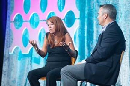 A man and a woman sitting on a stage engaged in a conversation. The woman is on the left side of the stage wearing a black dress and has long red hair. She is gesturing with her hands as if she is explaining something to the man on the right side. The man is wearing a suit and appears to be listening attentively. The background is a blue curtain with a pink and blue abstract design. The overall mood of the image is serious and focused.