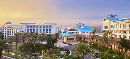 An aerial view of a resort at sunset. The sky is a beautiful orange and pink color with the sun setting in the background. The resort is surrounded by palm trees and other tropical plants. In the center of the image there is a large white building with a blue roof which appears to be a hotel or a casino. The building has multiple floors and balconies and there are several smaller buildings scattered throughout the resort. There is a fountain in the foreground and a few people can be seen walking around the area. The overall atmosphere of the resort is peaceful and serene.