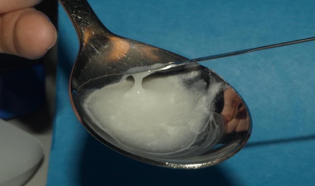 A hand holding a silver spoon with a small amount of white substance likely an opioid or another drug on it. The spoon is resting on a blue surface and the person's hand is visible on the left side of the image. A small scoop is being used to scoop or stir the substance. The background is blurred but it seems like the focus is on the spoon and the substance.