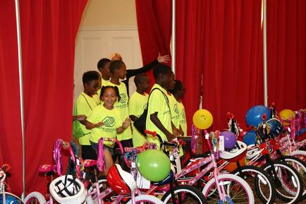A group of young children standing in front of a red curtain. They are all wearing yellow t-shirts with the logo of a sports team on them. The children are smiling and appear to be excited. In front of them there are several pink bicycles with colorful helmets and balloons attached to them. On the right side of the image there is a woman standing with her arms outstretched as if she is giving instructions or explaining something to the children. The background appears to be a room with a white door.