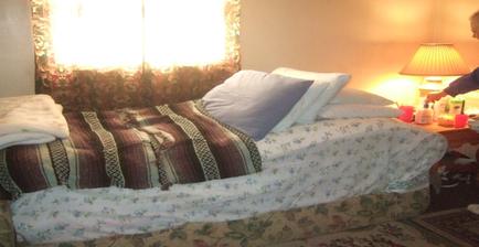 A bedroom with a single bed in the center. The bed is covered with a white comforter with a floral pattern and several pillows and blankets. On the right side of the bed there is a nightstand with a lamp and a few items on it. A woman is standing next to the nightstand looking at the bed. The room has a window with a curtain and a rug on the floor. The walls are painted in a light color and the overall atmosphere of the room is cozy and inviting.