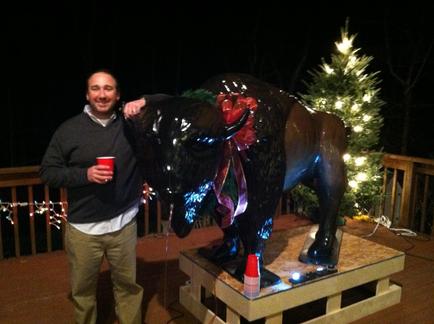 A man standing next to a large statue of a bison on a wooden platform. The bison is black and brown in color and is decorated with colorful lights and a red bow on its head. The man is holding a red cup in his left hand and is smiling at the camera. In the background there is a small Christmas tree with white lights and string lights. The platform appears to be on a deck or patio and the background is dark.