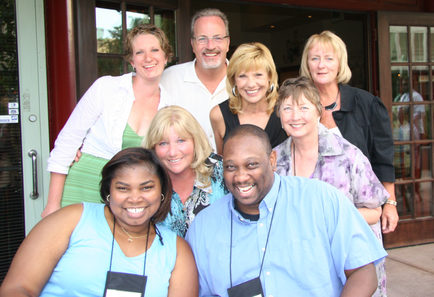 A group of nine people standing in front of a building with a glass door. There are nine people in total nine women and one man all of whom are smiling and posing for the photo. The group appears to be of different ages and ethnicity's and they are all dressed in casual clothing. The man in the center of the group is wearing a white shirt and glasses and he is standing with his arms around the woman on the left side of the image. The woman next to him is wearing an ID card around her neck. The other nine people are standing around her and all of them are looking at the camera. The background shows a brick building with large windows and a door.
