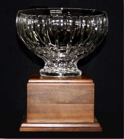 Of a glass bowl on a wooden base. The bowl is round and has a wide base with a narrow neck and a wide opening at the top. It appears to be made of clear glass with a ribbed pattern on the sides. The base is made of a dark wood with a smooth finish. The background is black making the bowl stand out. The overall appearance of the bowl is elegant and sophisticated.
