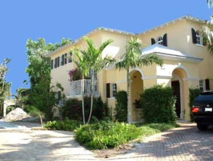 A two-story yellow house with black shutters and a balcony on the second floor. The house is surrounded by palm trees and other tropical plants. There is a driveway in front of the house and a black car parked on the right side. The sky is blue and the weather appears to be sunny and warm.