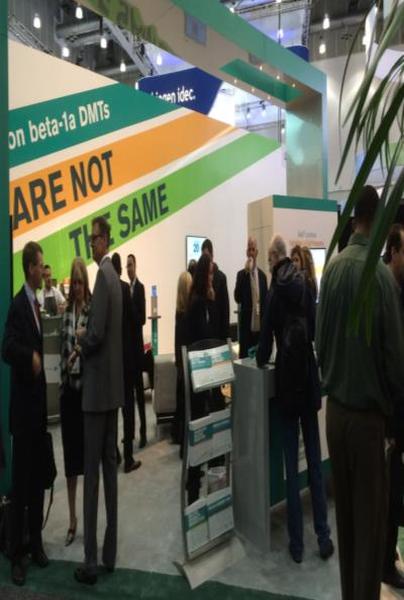 A group of people gathered around a booth at an exhibition. The booth has a green and white color scheme with the words "On beta-1a dots" written in bold letters. There is a sign that reads "Are not the same" in smaller letters below the sign. The people in the booth appear to be engaged in conversation and are looking at the information displayed on the booth. There are also some plants in the background suggesting that the exhibition is taking place indoors.