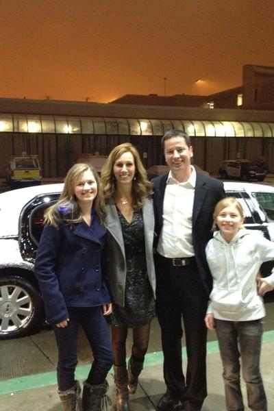 A family of four posing for a photo in front of a white car. The family consists of a man a woman and two young girls. The man is wearing a black suit and tie while the woman and girls are wearing casual clothing. The girl on the left is blonde and wearing a blue coat the girl in the middle is wearing black boots and the girl next to her is also blonde. They are all smiling and looking at the camera. In the background there is a building and a street lamp.