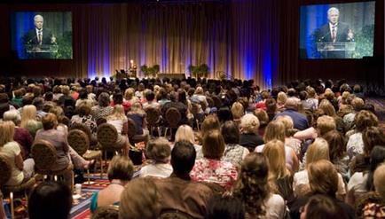 A large auditorium filled with people sitting in rows of chairs facing a stage. The stage is decorated with blue curtains and there are two large screens on either side of the stage displaying a man giving a speech. The audience appears to be attentively listening to the speaker. The people in the audience are of different ages and genders and they are all looking towards the stage with serious expressions on their faces. The overall mood of the image is one of concentration and focus.