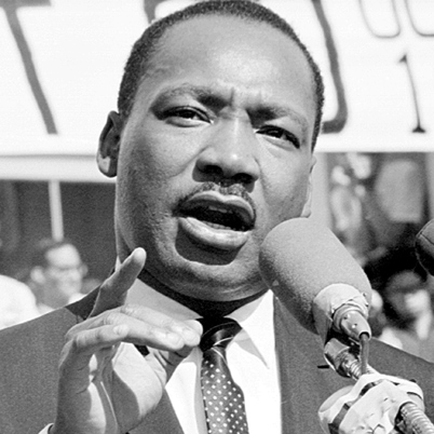 A black and white photograph of Martin Luther King Jr. speaking into a microphone. He is wearing a suit and tie and appears to be giving a speech. His mouth is open and his right hand is pointing upwards as if he is making a point. The background is blurred but it seems to be a crowd of people. The photograph is likely taken at a political rally or event.