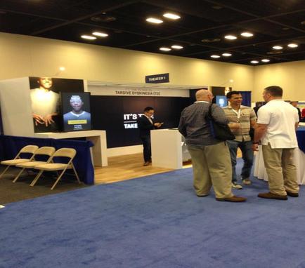 A trade show booth with a blue carpeted floor and white walls. There are several people gathered around the booth some of them are engaged in conversation. On the left side of the image there is a large screen displaying an image of a man in a suit and tie and on the right side there are several chairs and tables with white tablecloths. In the center of the booth is a sign that reads "It's time to take a break from the hustle and bustle of the trade show." The booth appears to be well-lit with bright lights.