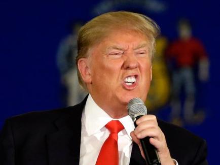 Former US President Donald Trump speaking into a microphone. He is wearing a black suit and a red tie. His mouth is open wide and his eyes are closed as if he is shouting or making a point. The background is blue with the seal of the United States of America on it. Trump appears to be giving a speech or addressing an audience.