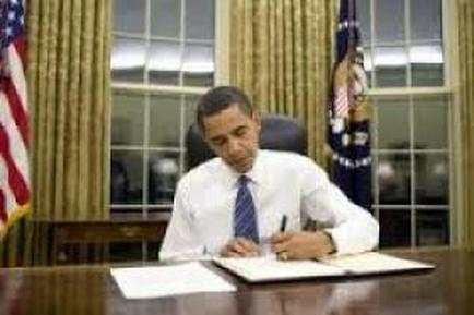 Former US President Barack Obama sitting at his desk in the Oval Office of the White House. He is wearing a white shirt and a blue tie and is holding a pen in his right hand. On the desk in front of him there is a large document with a pen resting on it. The desk is made of wood and there are two flags on either side of the desk. The background shows a large window with curtains and a flagpole with an American flag. The image appears to be taken during his signing of a document.